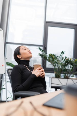 Asian designer holding coffee to go and relaxing near working table in office 