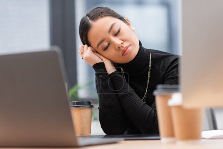 Foto de Sleepy asian designer sitting near computers and coffee in office - Imagen libre de derechos