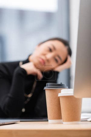 Foto de Coffee to go near computer and blurred interior designer in office - Imagen libre de derechos