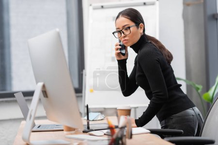 Asian designer in eyeglasses looking at computer and talking on smartphone in studio 