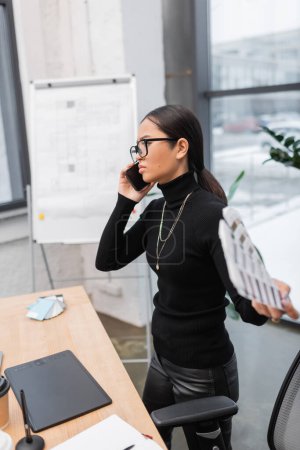 Foto de Asian designer in eyeglasses talking on smartphone and holding color swatches in office - Imagen libre de derechos