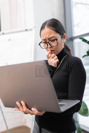 Photo for Stressed asian designer holding blurred laptop in studio - Royalty Free Image