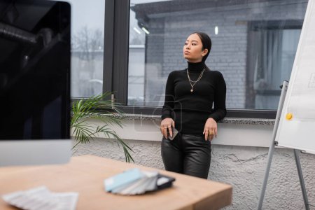 Asian designer holding smartphone near window in office 