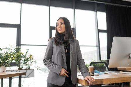 Brunette asian designer in jacket standing near working table in office 