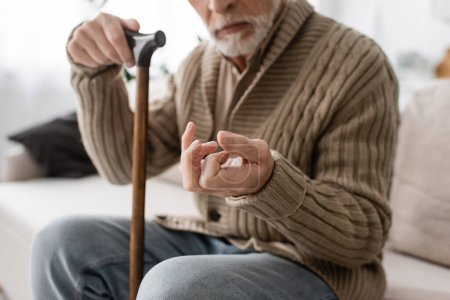 cropped view of senior man with parkinson disease holding walking cane while sitting on couch at home