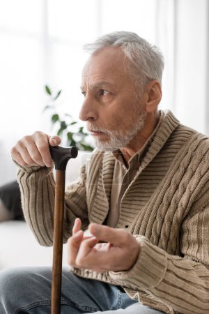 Foto de Depressed man with parkinsonian syndrome sitting with walking cane and looking away at home - Imagen libre de derechos