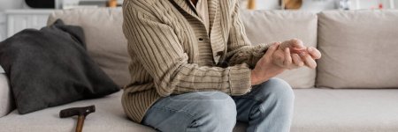 Foto de Partial view of aged man with trembling hands suffering from parkinsonian syndrome while sitting on sofa, banner - Imagen libre de derechos