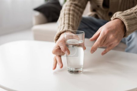 partial view of aged man with parkinson disease and trembling hands taking glass of water from table at home