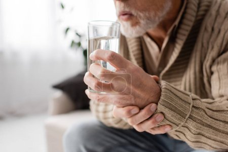 cropped view of aged man with parkinsonism holding glass of water in trembling hands while sitting at home