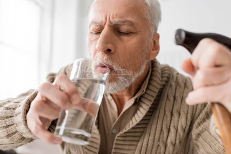 Foto de Senior man with parkinson disease and trembling hands holding glass of water and walking cane on blurred foreground - Imagen libre de derechos