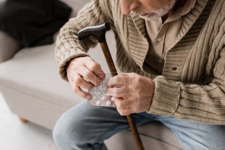 Foto de Cropped view of senior man with parkinson disease sitting with walking cane and pills on couch at home - Imagen libre de derechos