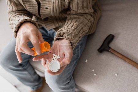 Foto de Cropped view of aged man suffering from parkinsonism and holding container with pills while sitting on couch - Imagen libre de derechos