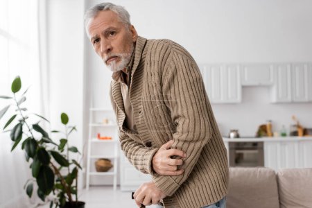 Foto de Grey haired man with parkinson disease standing with walking cane in kitchen - Imagen libre de derechos