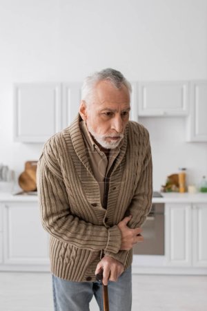 depressed senior man with parkinson disease standing with walking cane in kitchen