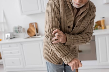 partial view of senior man with parkinson disease and hands tremor standing with walking cane in kitchen