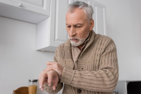 grey haired man with parkinson disease looking at trembling hands in kitchen