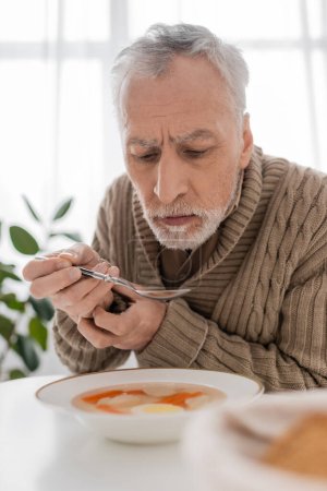 Foto de Senior man with parkinson disease holding spoon in trembling hands near plate with soup in kitchen - Imagen libre de derechos
