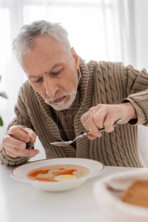 Foto de Aged man with parkinson disease and hands tremor sitting with spoon near plate with soup in kitchen - Imagen libre de derechos