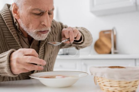 Foto de Aged man with parkinson disease holding spoon in trembling hand while eating soup in kitchen - Imagen libre de derechos