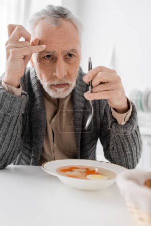 depressed man with parkinsonian syndrome looking away while holding spoon near soup in kitchen