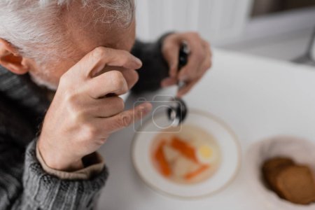 Photo for High angle view of man with parkinson syndrome and trembling hands holding spoon near blurred soup in kitchen - Royalty Free Image