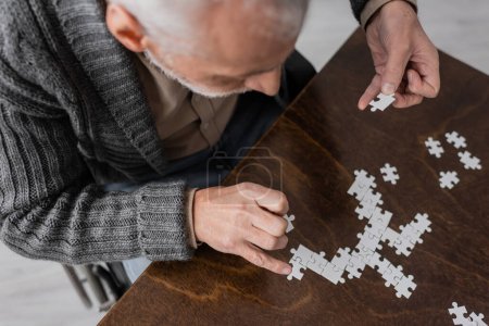 overhead view of senior man with parkinson disease and tremor in hands combining elements of jigsaw puzzle on table at home