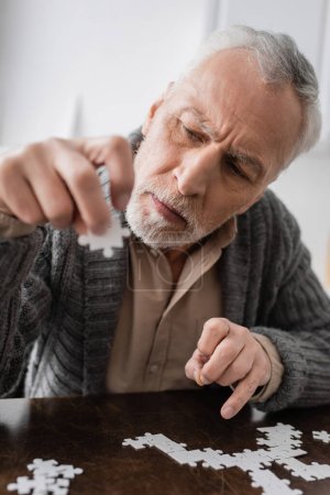 Foto de Grey haired man suffering from parkinsonian syndrome and holding element of jigsaw puzzle in trembling hand - Imagen libre de derechos