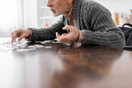 Foto de Cropped view of man with parkinson disease and trembling hands sitting near jigsaw puzzle at home - Imagen libre de derechos