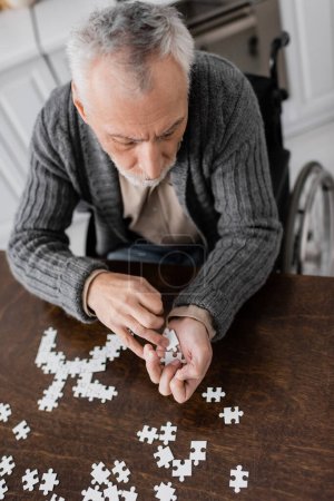 high angle view of man with parkinsonian syndrome sitting in wheelchair and holding elements of jigsaw puzzle in trembling hands