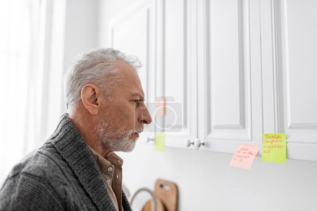 Téléchargez les photos : Side view of grey haired man with alzheimer syndrome looking at sticky notes with names and phone numbers in kitchen - en image libre de droit
