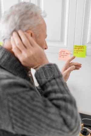 Photo for Blurred man suffering from memory loss and pointing at sticky notes with names and phone numbers in kitchen - Royalty Free Image