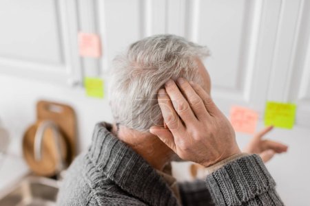 Foto de Grey haired senior man with alzheimer syndrome touching head and pointing with blurred sticky note in kitchen - Imagen libre de derechos