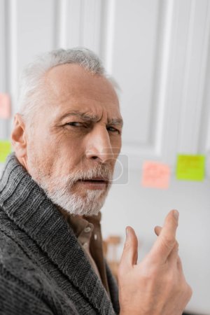 Foto de Thoughtful man with alzheimer disease looking at camera near blurred sticky notes in kitchen - Imagen libre de derechos