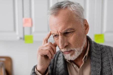 thoughtful senior man with alzheimer disease touching head near blurred sticky notes in kitchen