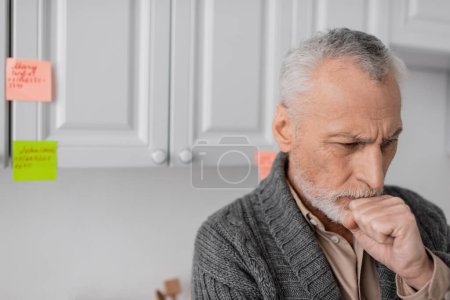 Foto de Aged man with alzheimer syndrome holding fist near face while thinking near blurred sticky notes in kitchen - Imagen libre de derechos