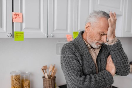 depressed man with alzheimer disease touching forehead while standing near sticky notes in kitchen