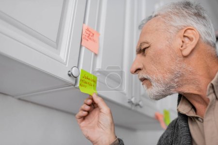 Foto de Senior man with alzheimer syndrome looking at sticky note with name and phone number in kitchen - Imagen libre de derechos