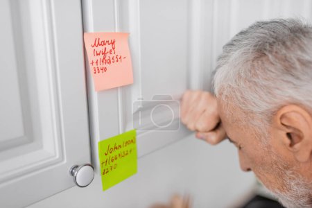 stressed man suffering from memory loss and standing near sticky notes with names and phone numbers in kitchen