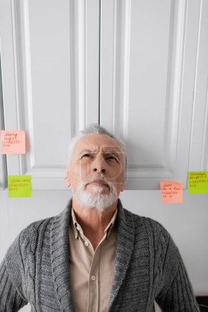 Téléchargez les photos : Senior man suffering from memory loss and looking up while standing near sticky notes with names and phone numbers in kitchen - en image libre de droit