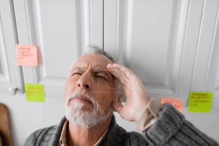 Foto de Senior man with closed eyes and hand near head standing near blurred sticky notes while suffering from memory loss - Imagen libre de derechos