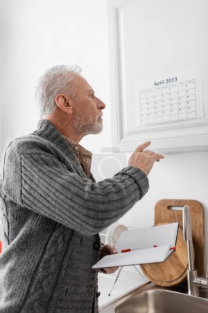 Foto de Senior man with alzheimer disease holding blank notebook and pointing at calendar in kitchen - Imagen libre de derechos