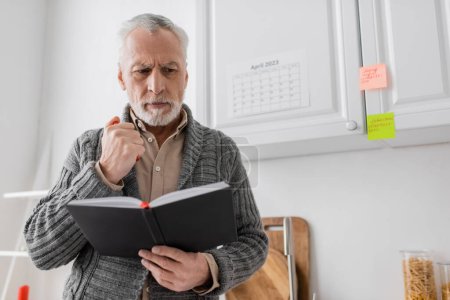 thoughtful man suffering from memory loss and standing with notebook and felt pen in kitchen