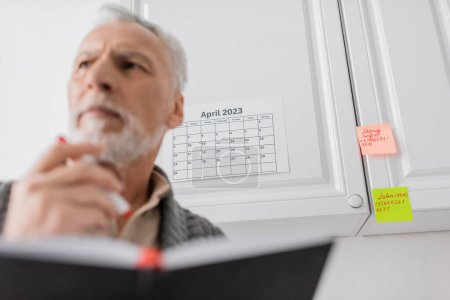 Foto de Low angle view of thoughtful man suffering from memory loss and standing with blurred notebook near calendar and sticky notes - Imagen libre de derechos