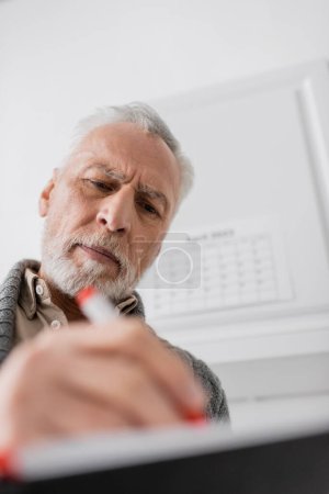 low angle view of bearded senior man with alzheimer disease writing in blurred notebook at home