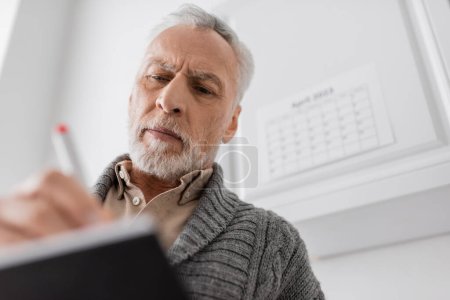 low angle view of grey haired man with alzheimer syndrome writing in blurred notebook