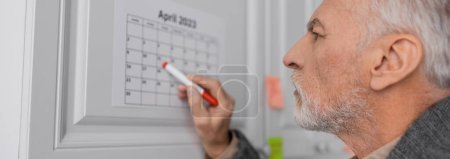 Foto de Side view of senior man with alzheimer disease pointing with felt pen at calendar in kitchen, banner - Imagen libre de derechos