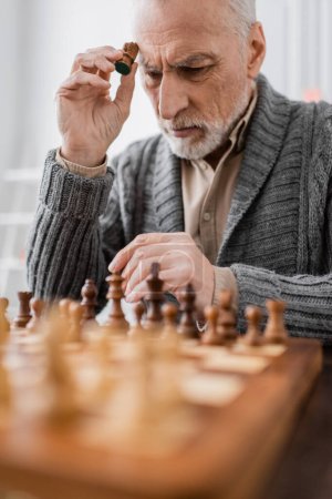 aged man sick on alzheimer syndrome holding figure while thinking near chessboard on blurred foreground