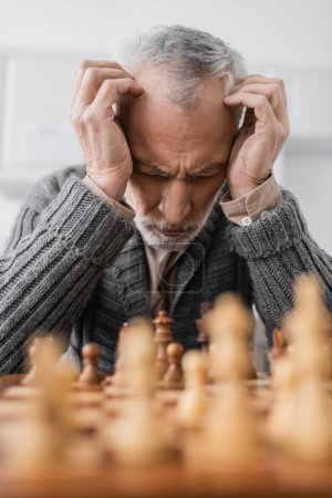 Foto de Depressed man with alzheimer disease sitting with closed eyes and hands near head at chessboard on blurred foreground - Imagen libre de derechos
