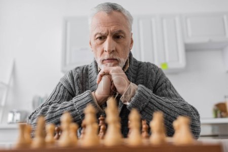 senior man suffering from memory loss and thinking near blurred chessboard at home