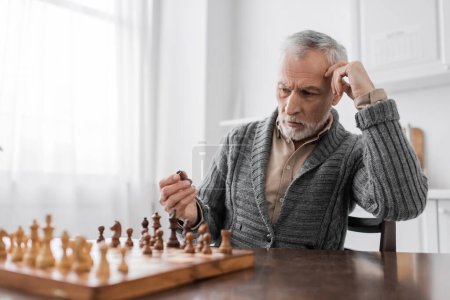 grey haired man with alzheimer disease holding chess figure and thinking at home